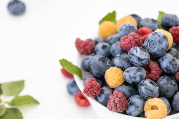 Blueberry and raspberry is tasty, healthy and delicious fruits. Fresh blueberries with red and yellow raspberries with leaves of mint in white bowl. Wild berry in white plate on white background.