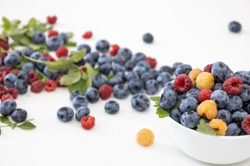 Blueberry and raspberry is tasty, healthy and delicious fruits. Fresh blueberries with red and yellow raspberries with leaves of mint in white bowl. Wild berry in white plate on white background.