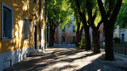 Obraz premium Picturesque Cobblestone Street with Yellow Building, Lush Trees, and Historic Brick Background for Travel Magazines, Heritage Projects, or European-Themed Content.