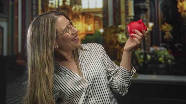Middle aged blonde woman smiling and holding red piggy bank in church building interior; charity faith joy.