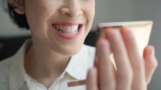 Young Asian woman in a well-lit space. She is captured in the act of carefully putting a dental retainer into her mouth, her eyes fixed on her reflection in a small, handheld mirror.