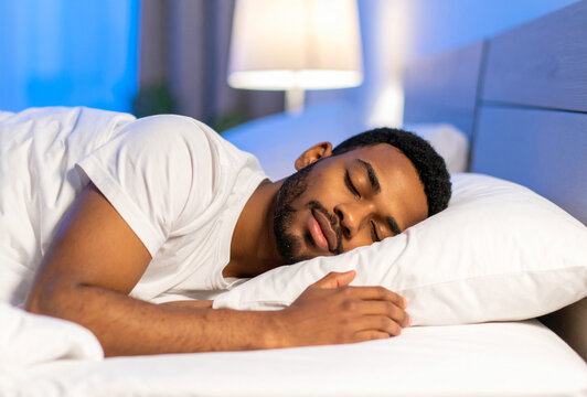Un hombre joven dormido descansando durante la noche en una cama con sabanas y almohada blanca con la luz del dormitorio encendida