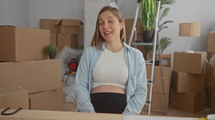Woman with pregnant belly waves at camera in new apartment interior; anticipation joy.