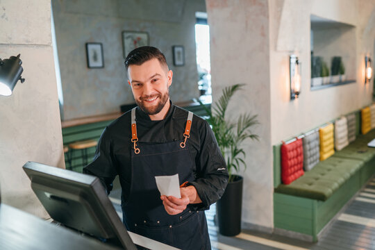 Stylish bearded smiling waiter dressed black uniform processing customer orders using point of sale order terminal system touch screen. Successful people teamwork, restaurant industry concept image. - Powered by Adobe