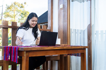 A young Asian woman in a white t-shirt works on a laptop at a wooden desk outdoors, writing notes and smiling. Remote work, freelance lifestyle, digital nomad, and online learning concept.