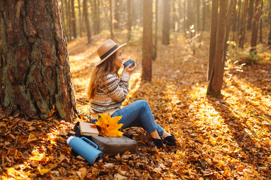 Happy woman in hat pouring tea from blue thermos into cup while sitting by tree in autumn forest. Young woman enjoying adventure in nature, drinking tea and having fun in sunset rays. Active lifestyle