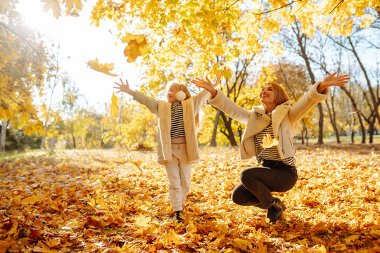 Happy mother and daughter having fun in an autumn park at sunset. Young woman with her beloved daughter throwing autumn leaves up and spending time together outdoors. Fun and childhood concept.