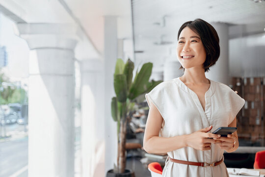 Asian mature business woman ceo investor using cell phone mobile app. Portrait of eastern female businesswoman holding smartphone in office working for trading, looking dreaming aside. Copy space - Powered by Adobe