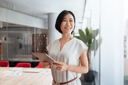 Eastern Asian 40s adult manager business woman looking aside dreaming, holding digital touchscreen device. Mature CEO businesswoman using tablet using fintech tab application in office. Copy space