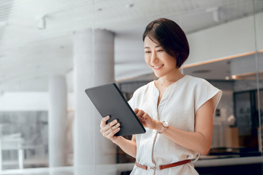 Mature CEO businesswoman holding digital tablet using fintech tab application standing in office. Eastern Asian 40s adult professional business woman looking digital touchscreen device. Copy space