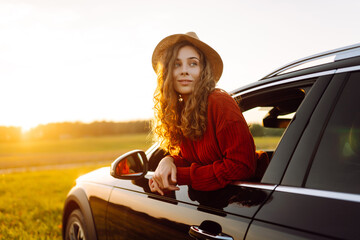 Female traveler in hat leans out of car window, enjoying sunset against the backdrop of peaceful...