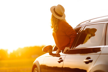 Female traveler in hat leans out of car window, enjoying sunset against the backdrop of peaceful...