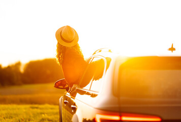 Female traveler in hat leans out of car window, enjoying sunset against the backdrop of peaceful...