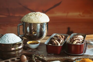 A vintage kitchen setting shows a mixing bowl with rising dough, awaiting baking in loaf pans, with flour, an egg, and other baking ingredients surrounding it. The background suggests sunset colors