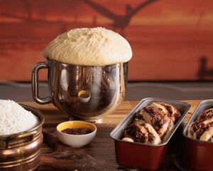 Fresh dough rises enthusiastically in a metal mixing bowl, near ingredients and loaves in baking...