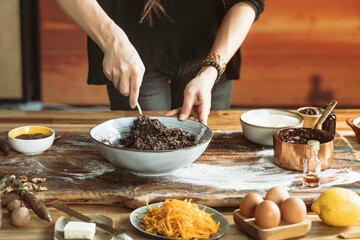 A person mixes dark, rich ingredients in a bowl on a rustic wooden surface. Other ingredients like eggs, carrots, lemon, and butter await their turn in this gourmet preparation