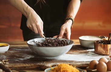 With focused attention, a person mixes poppy seeds in a bowl on a flour-dusted wooden table. Eggs, shredded carrots, and other ingredients are carefully arranged nearby