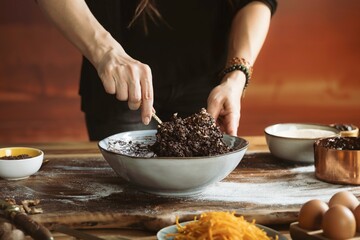 Hands are mixing a dense, dark poppy seed filling in a grey bowl. Several ingredients such as eggs, shredded carrots and sugar surround the bowl in a warm and inviting setting
