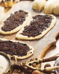 Several flattened dough pieces are shown covered with a dark, rich poppy seed filling on a worn wooden board. A nutcracker, walnuts, and other ingredients lie nearby