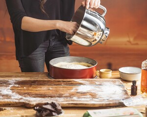 A woman is pouring cake batter from a stainless steel mixing bowl into a red cake pan, on a countertop covered in flour while baking in her kitchen