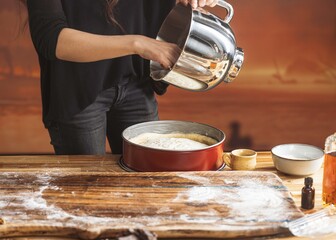 A baker in black pours batter from a silver mixing bowl into a red cake pan. This happens on a flour-dusted wooden table during golden hour. Spices and sugar sit near