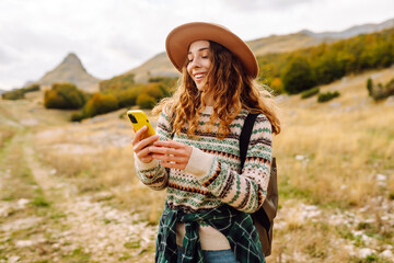 Beautiful woman in cozy sweater and hat uses smartphone against backdrop of mountain landscape....