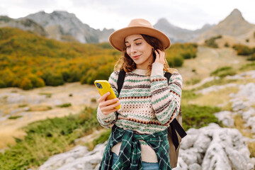 Beautiful woman in cozy sweater and hat uses smartphone against backdrop of mountain landscape....