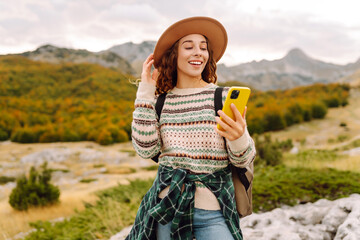 Beautiful woman in cozy sweater and hat uses smartphone against backdrop of mountain landscape....