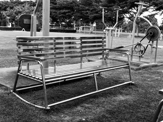 A grayscale photograph of a sleek, shiny metal bench located within an outdoor fitness park. The...