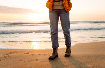 Close-up of a foot in hiking boots standing on a sandy sunset beach. A female traveler in boots in...