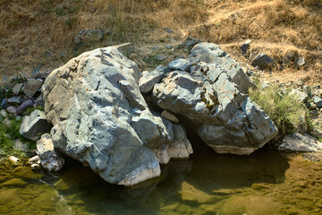 Rock face with moss and lichens by the water’s edge of a mountain stream in Boshkizilsay, Uzbekistan. Sunny summer day, view of the natural shore and water reflection