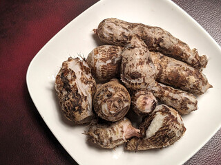 A close-up view of a white plate filled with raw, unpeeled taro roots showcasing their textured brown skin and white flesh with a dark red textured background