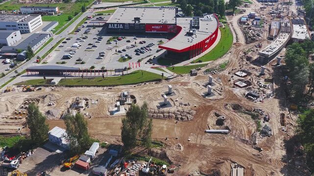 Large scale road construction with excavators, drilling equipment, a red building labeled Aleja, a communication tower, and workers in motion.