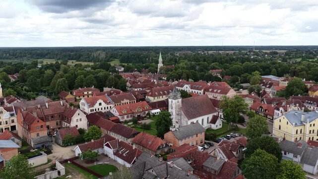 Scenic aerial panorama of Kuldiga&rsquo;s old town in Latvia, showcasing the white church with a clock tower, red-tiled rooftops, and lush greenery under a cloudy summer sky.