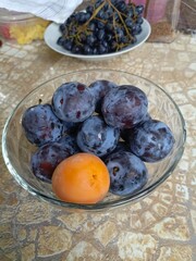 Plums and an apricot in a glass bowl in a realistic still life style in purple, orange, and beige colors, representing healthy eating and harvest
