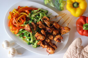 Grilled chicken skewers served with stir fried red and yellow capsicum and broccoli on a white plate