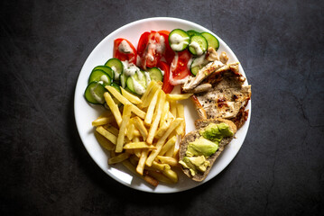 Healthy plate: grilled chicken, salad, avocado, and fries on dark background.