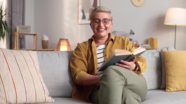 Creative pretty woman in glasses holding paper book, looking at camera and smiling. Beautiful female sitting on couch in living room, relaxing and reading. Lifestyle and leisure time.