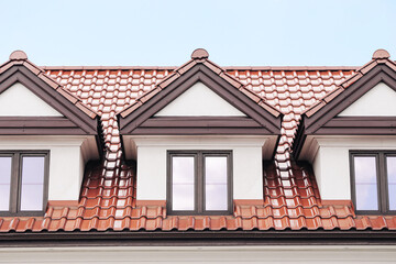 Three dormer windows on modern house roof. Red ceramic tiles under blue sky. White facade with brown trim around glass panes. Symmetrical architecture detail in daylight. Residential building rooftop 