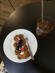 Danish pastry with fresh berries and cream on a white plate with fork, served with a glass of iced coffee on a black table, top view, fresh bakery and summer drink in a cafe