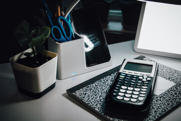 an evening desk set up for studying or work with a calculator, notebook, lamp, and a smartphone docked in a charging stand. back to school or college design element