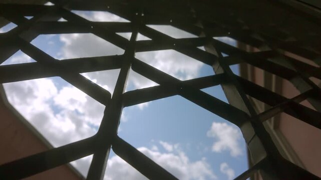 Geometric security grill with crisscross metal pattern frames bright blue sky and clouds viewed from below at building entrance