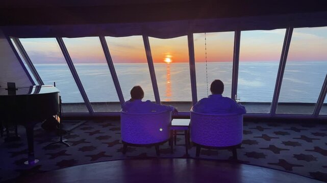 Peaceful 4K footage of a couple relaxing aboard a cruise ship while watching a vibrant sunset over the open ocean. Warm evening light reflects across the water through panoramic windows, creating a ro