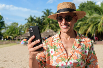 Hispanic Male Tourist in Panama Hat and Tropical Shirt Summer Vacation at Beach Using Phone