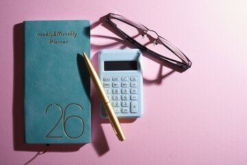 2026 small planner calculator, eyeglasses and pen against pink background.