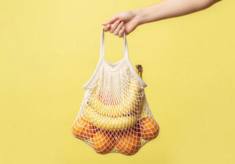 A hand holds a white, reusable mesh grocery bag filled with fresh bananas and oranges against a bright yellow background, promoting an eco-friendly and healthy lifestyle.