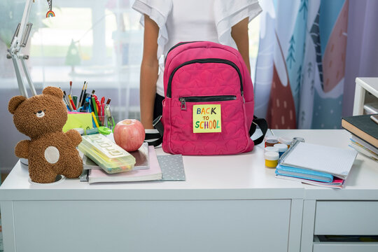 A pink school backpack with back to school text stands on a table filled with stationery items. Nearby are an apple and a teddy bear, creating a cheerful study environment.