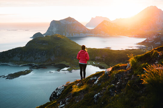 Young woman hiker standing on a rocky cliff in Lofoten Islands, Norway, during sunrise in summer, with the golden midnight sun lighting the fjords. - Powered by Adobe