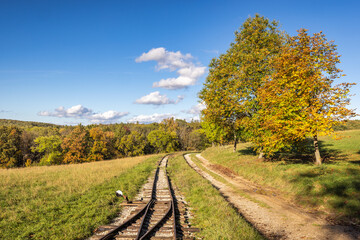 Railroad tracks wind through an idyllic autumn landscape under a clear blue sky with scattered clouds.