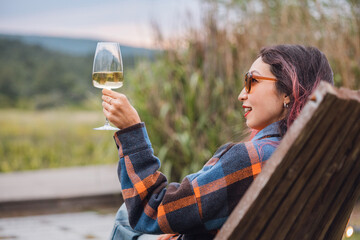 Young woman sitting on wooden chair, holding glass of white wine, enjoying landscape in vineyard